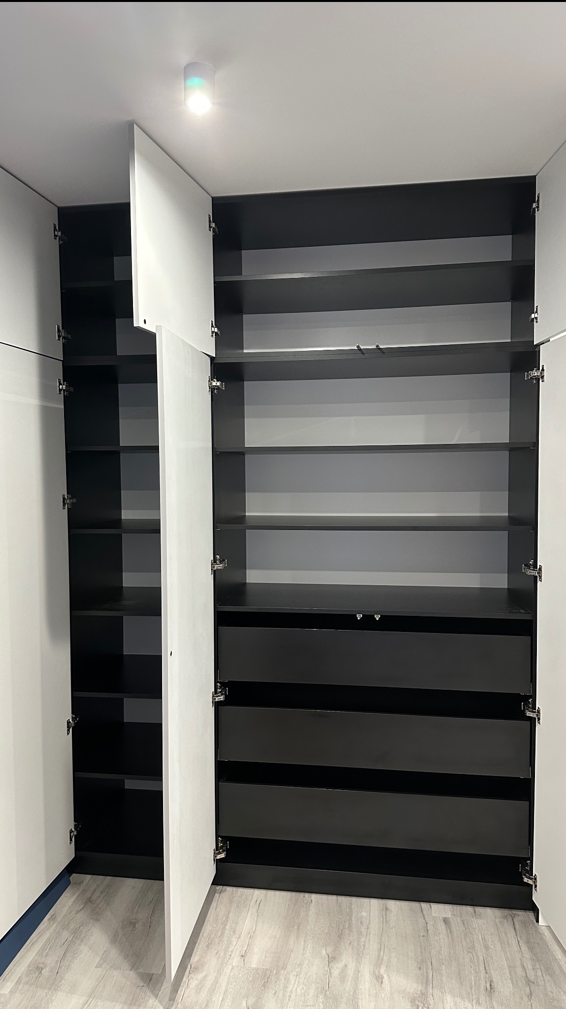 Nowoczesna szafa wnękowa z otwartymi drzwiami, ukazująca czarne półki i szuflady na tle białych frontów. Minimalistyczny design, oświetlenie punktowe.