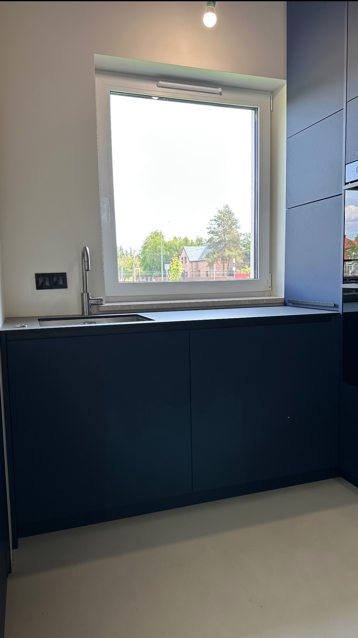 Nowoczesna, granatowa zabudowa kuchenna pod oknem z widokiem na budynek. Minimalistyczny design, gładkie fronty, metalowa bateria. Jasna podłoga.