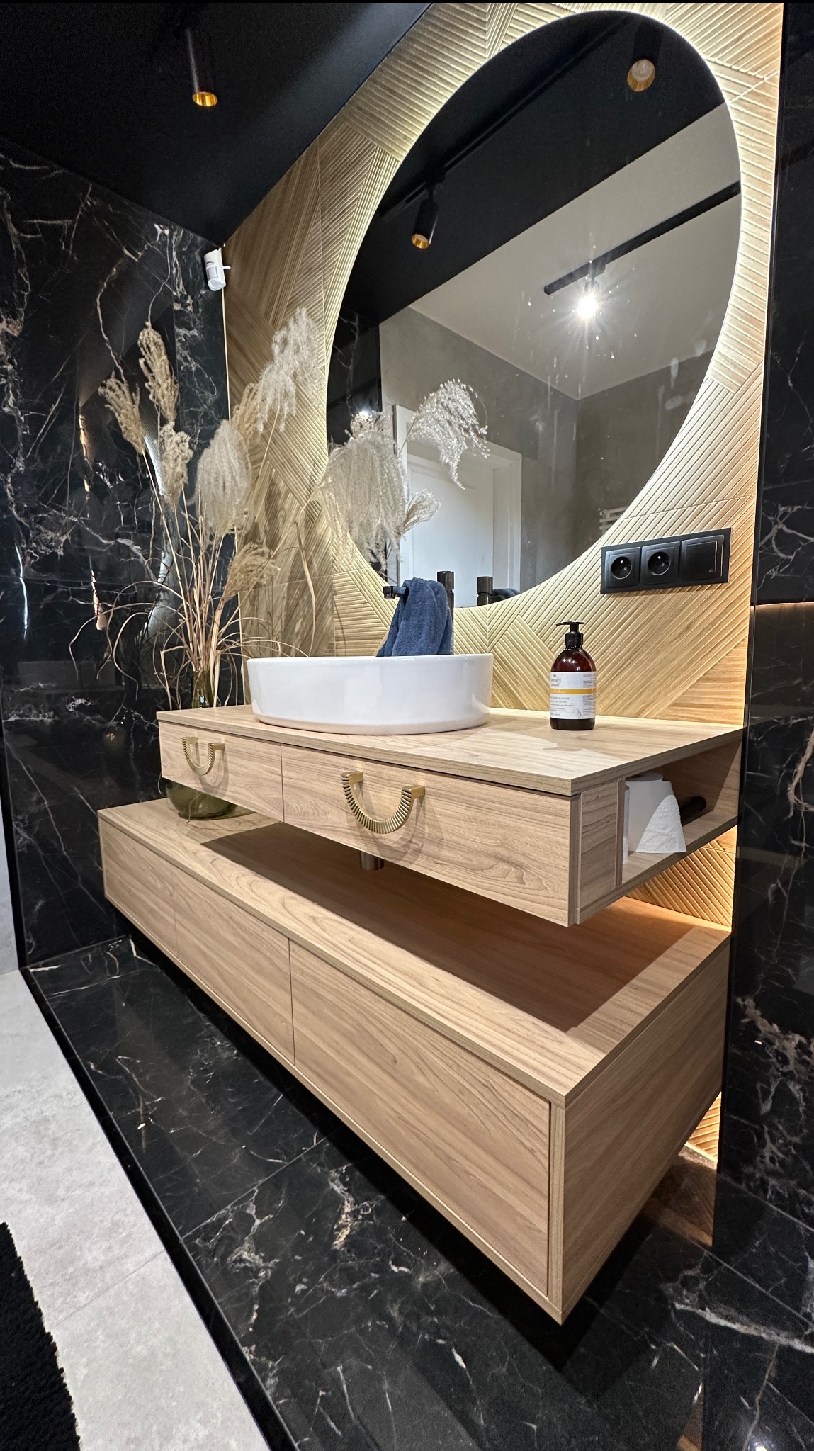 Nowoczesna szafka łazienkowa z drewnianymi frontami, okrągłe lustro z podświetleniem LED, czarne marmurowe ściany i podłoga. Umywalka nablatowa.