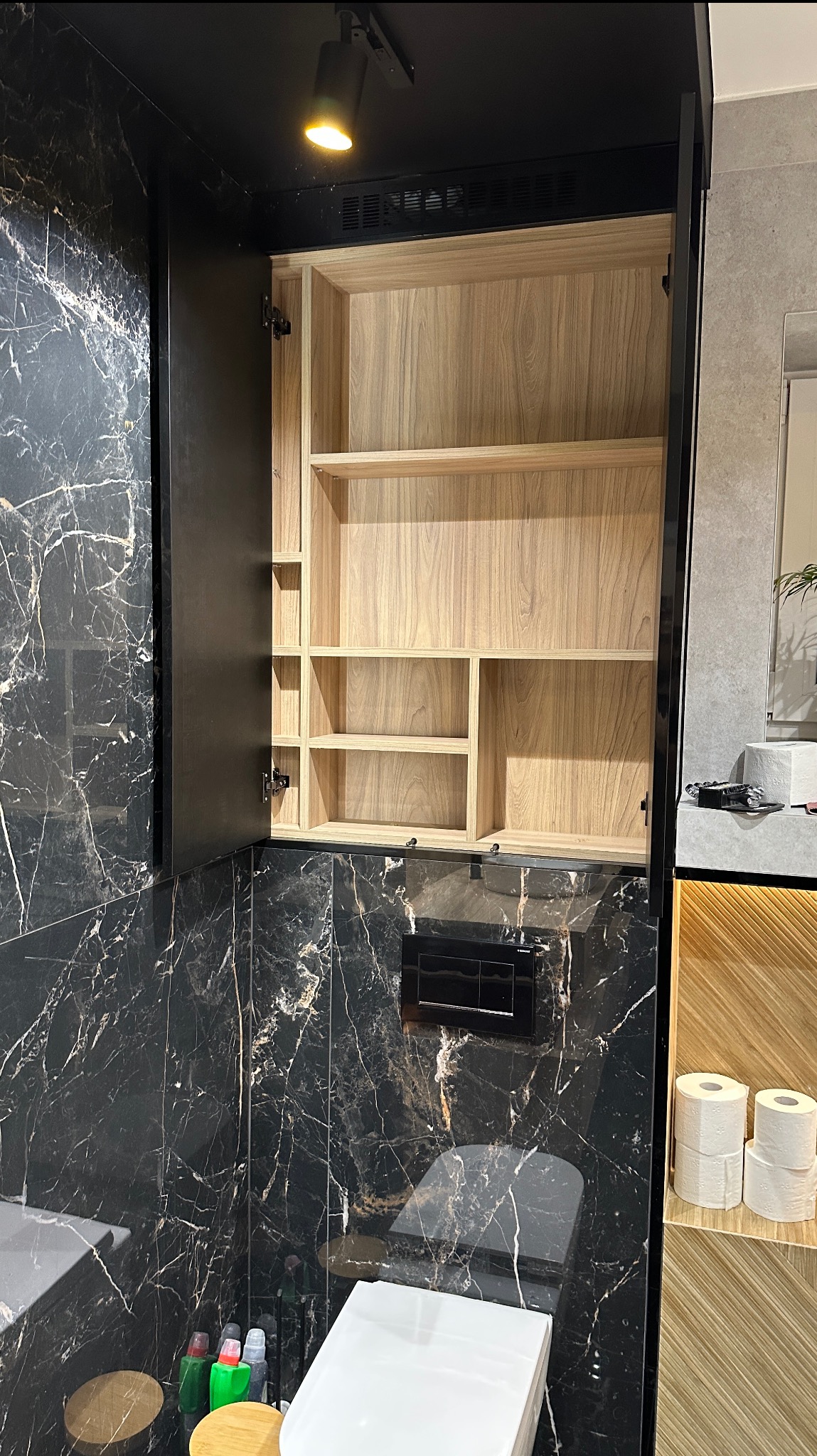Otwarta szafka łazienkowa z drewnianymi półkami na tle ściany z czarnego marmuru. Widoczny fragment toalety i umywalki. Oświetlenie punktowe.