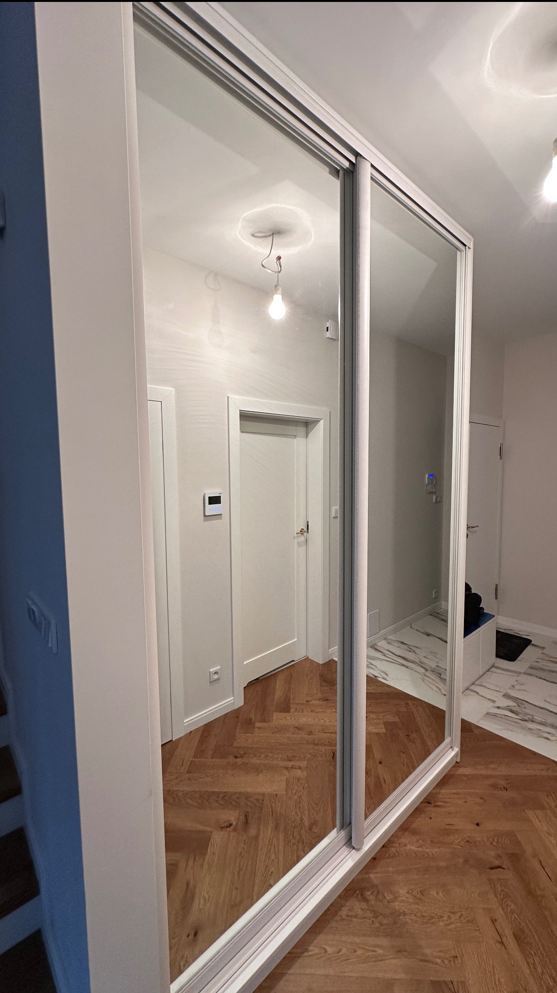 Nowoczesna szafa wnękowa z lustrzanymi drzwiami przesuwnymi, odbijająca parkiet w jodełkę i białe drzwi w mieszkaniu w Warszawie. Minimalistyczny design.