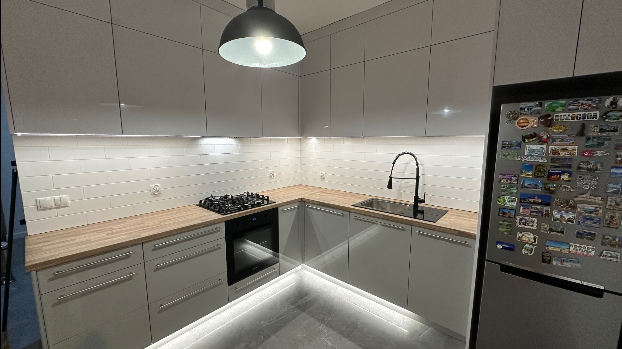 Jasnoszara kuchnia w kształcie litery L z drewnianym blatem, oświetleniem LED pod szafkami i czarną armaturą, uzupełniona o lodówkę z magnesami.