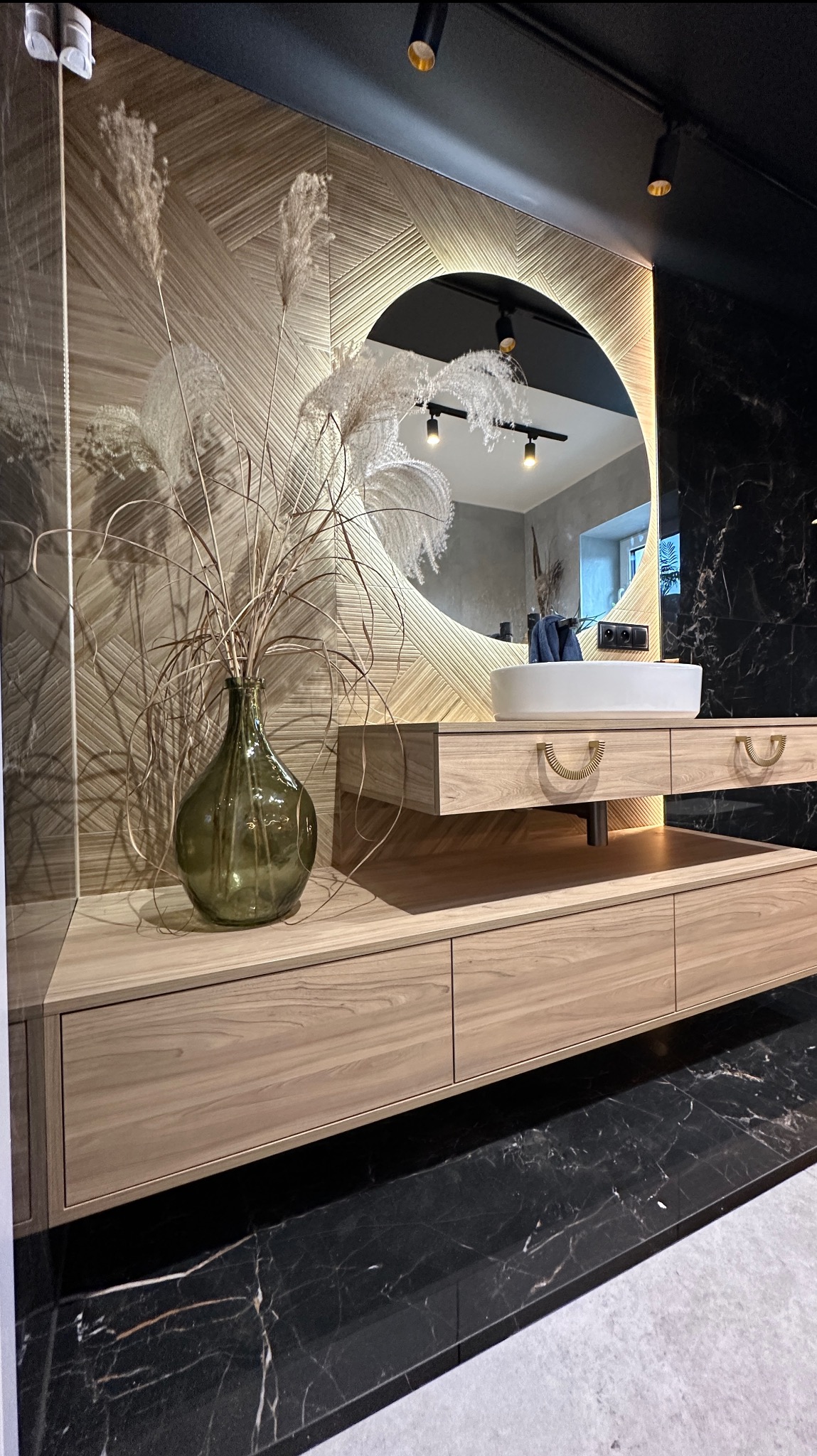 Nowoczesna łazienka z drewnianą szafką pod umywalkę, okrągłym lustrem z podświetleniem LED i marmurową podłogą. Wazon z trawami pampasowymi dodaje naturalnego akcentu.
