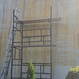 BADI Robert Badecki - Elewacja budynku z widocznymi zabrudzeniami, rusztowanie metalowe z drewnianą platformą, drabina oparta o rusztowanie, oraz ozdobne krzewy iglaste u podstawy elewacji.