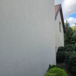 Robert Badecki - Teksturowana, jasna elewacja budynku jednorodzinnego z widocznym fragmentem dachu z brązową dachówką, fragmentem okna i kamerą monitoringu. Zza rogu budynku wystaje głowa mężczyzny w niebieskiej...