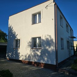 Robert Badecki - Odnowiona, kremowa elewacja domu jednorodzinnego z brązowymi rynnami i podmurówką, widoczne rusztowanie z boku budynku, cień drzewa na ścianie.