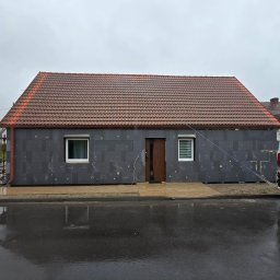 Robert Badecki - Dom z szarą elewacją w trakcie ocieplania, widoczne płyty izolacyjne. Czerwony dach, mokry chodnik i ulica po deszczu. Ujęcie z poziomu ulicy.