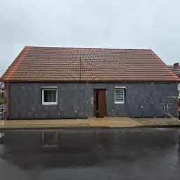 Dom z szarą elewacją w trakcie ocieplania, widoczne płyty izolacyjne. Czerwony dach, mokry chodnik i ulica po deszczu. Ujęcie z poziomu ulicy.