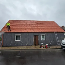 Czyszczenie elewacji domu z szarego styropianu przez pracownika w żółtej kamizelce za pomocą myjki ciśnieniowej. Widoczne maszyny czyszczące i drabina.