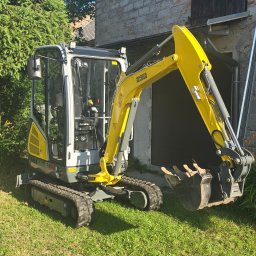 BADI Robert Badecki - Żółto-szara mini koparka Wacker Neuson na trawie, z budynkiem w tle. Widoczne detale kabiny i łyżki. Sprzęt gotowy do pracy.