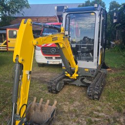 BADI Robert Badecki - Żółto-szara mini koparka Wacker Neuson na trawiastym terenie, z widocznym łyżką i gąsienicami. W tle budynek z czerwonej cegły i biały bus z czerwonym pasem.