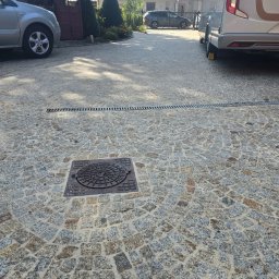 BADI Robert Badecki - Ułożona kostka brukowa w odcieniach szarości i brązu z widoczną studzienką kanalizacyjną i liniowym odpływem. W tle dom, samochody i przyczepa kempingowa.
