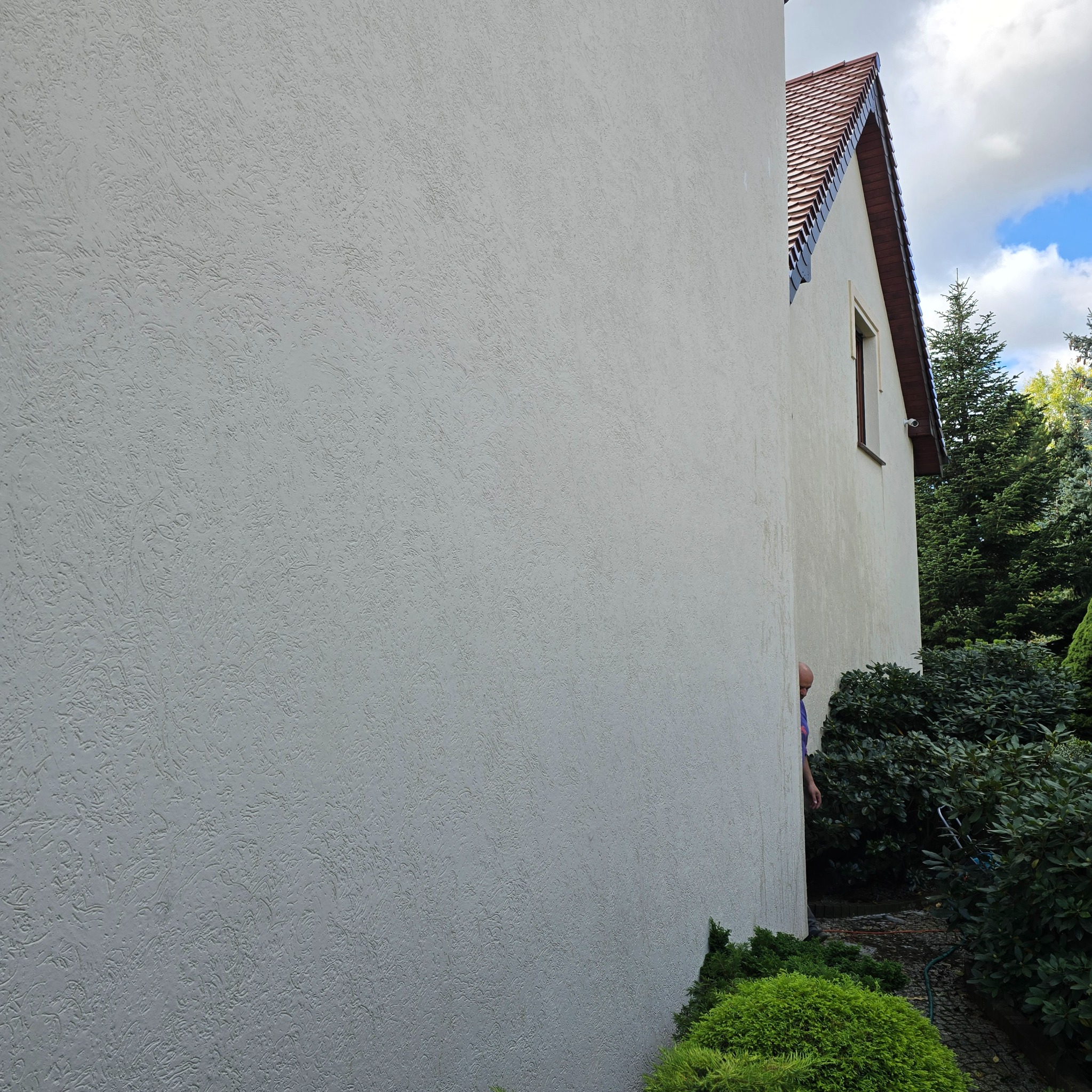 Teksturowana, jasna elewacja budynku jednorodzinnego z widocznym fragmentem dachu z brązową dachówką, fragmentem okna i kamerą monitoringu. Zza rogu budynku wystaje głowa mężczyzny w niebieskiej...