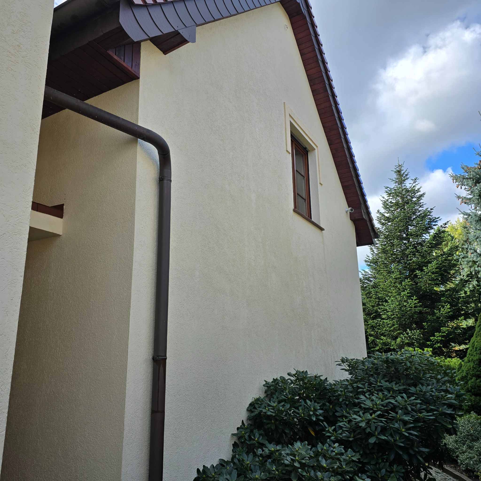 Jasna elewacja domu jednorodzinnego z widocznym oknem, rynną i fragmentem dachu z brązową dachówką, otoczona zielenią