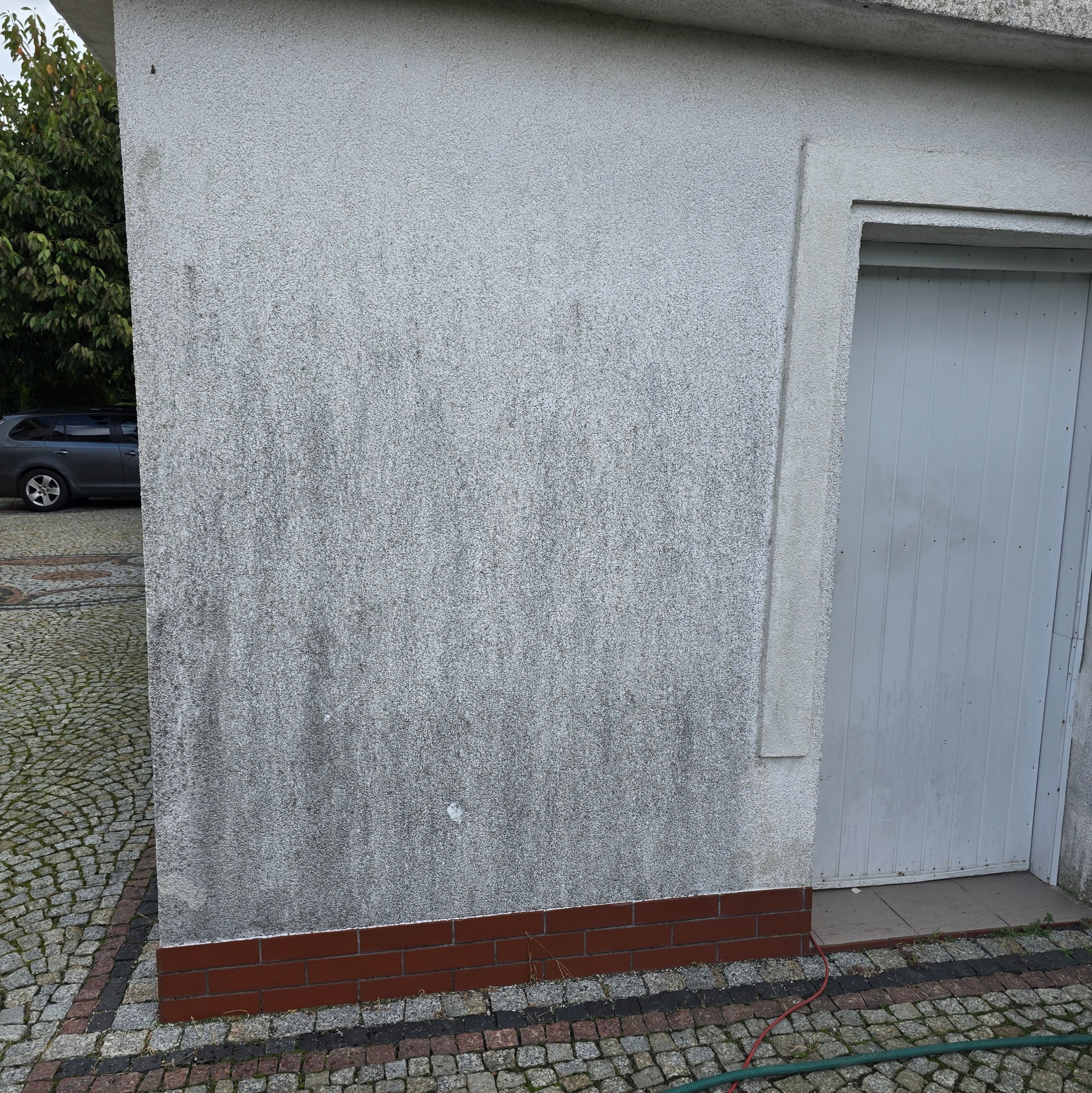 Szara, brudna elewacja budynku z widocznymi zaciekami i zabrudzeniami, z czerwoną ceglaną podmurówką i szarą bramą garażową po prawej stronie, na tle kostki brukowej i zaparkowanego samochodu.