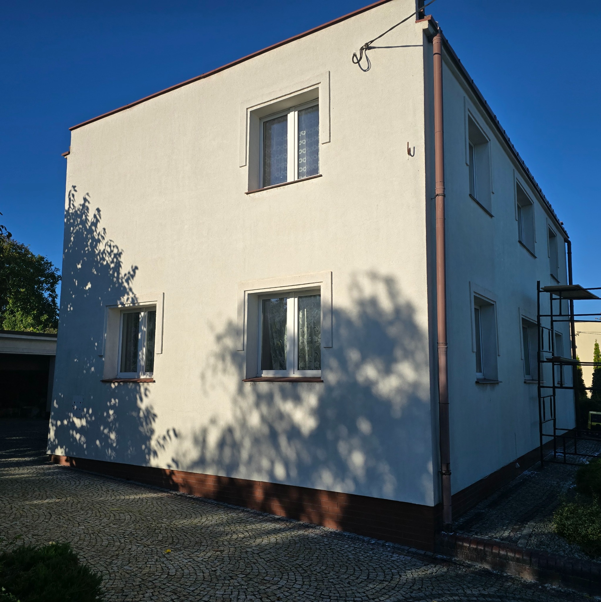 Odnowiona, kremowa elewacja domu jednorodzinnego z brązowymi rynnami i podmurówką, widoczne rusztowanie z boku budynku, cień drzewa na ścianie.