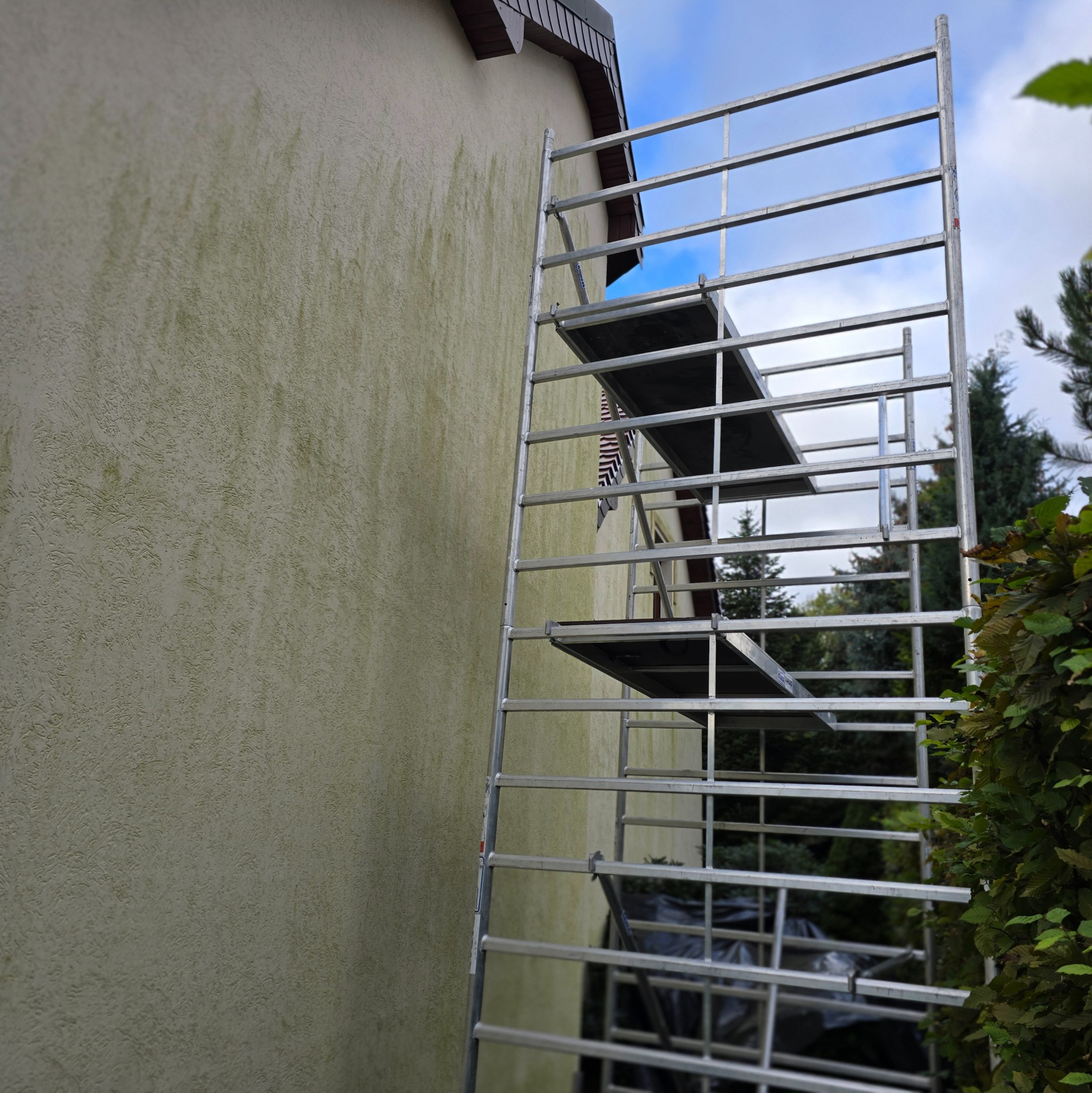 Metalowe rusztowanie stojące przy ścianie budynku z zielonkawym, chropowatym tynkiem, widoczne ślady zabrudzeń i porostów, w tle zieleń drzew i krzewów.