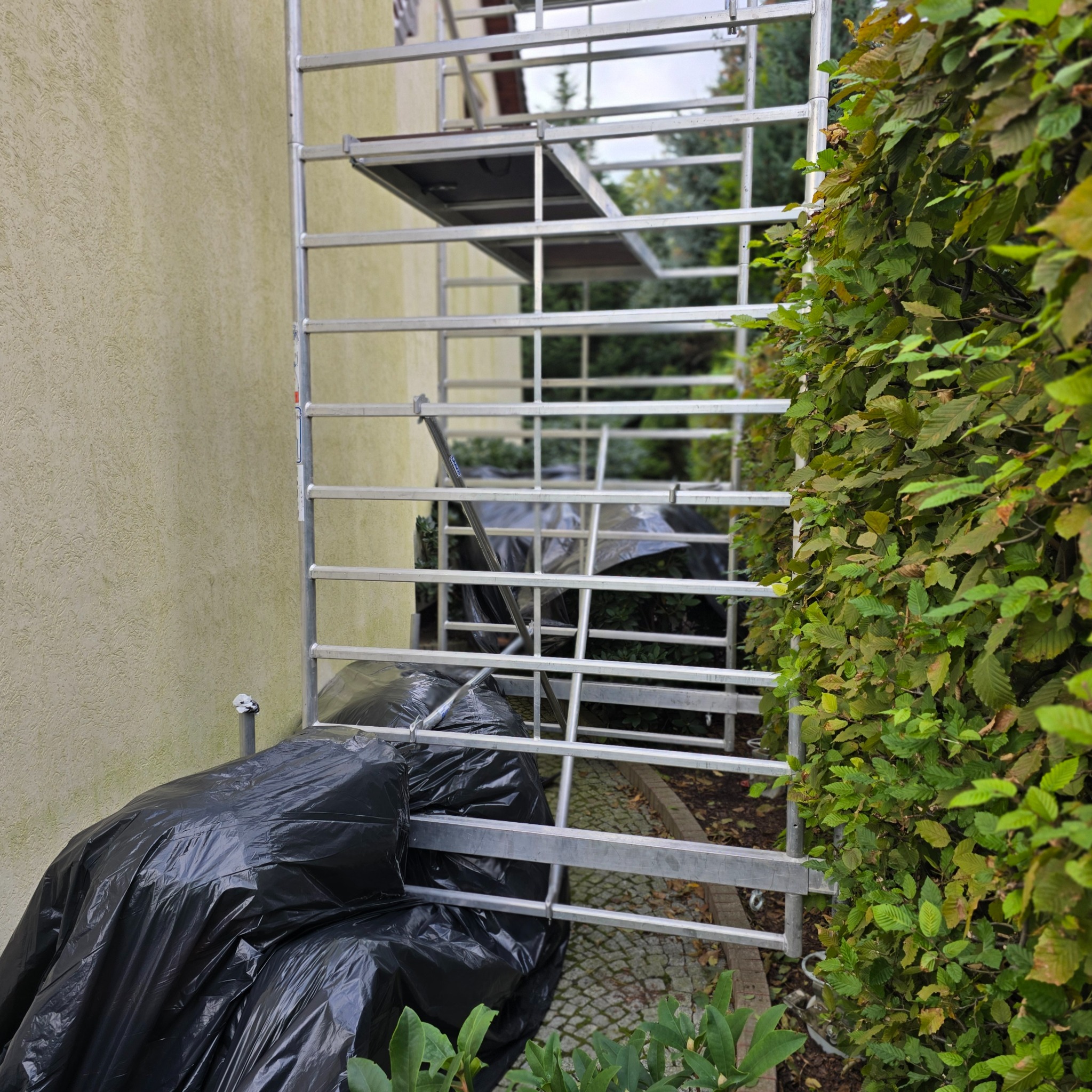 Metalowe rusztowanie ustawione przy ścianie budynku z fakturą baranka, zabezpieczone czarną folią, z widocznym fragmentem żywopłotu.