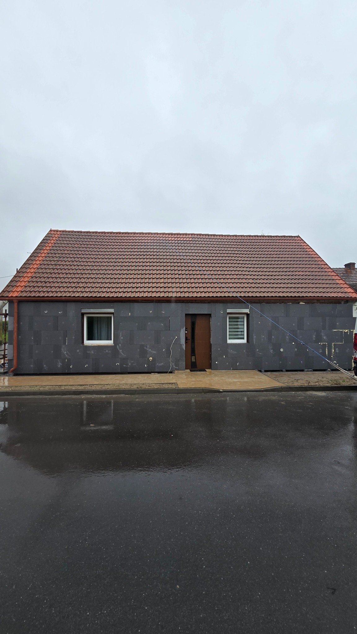 Dom z szarą elewacją w trakcie ocieplania, widoczne płyty izolacyjne. Czerwony dach, mokry chodnik i ulica po deszczu. Ujęcie z poziomu ulicy.