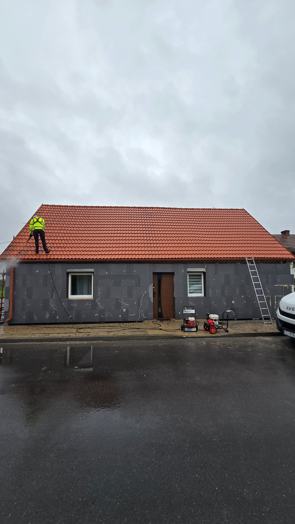 Czyszczenie elewacji domu z szarego styropianu przez pracownika w żółtej kamizelce za pomocą myjki ciśnieniowej. Widoczne maszyny czyszczące i drabina.