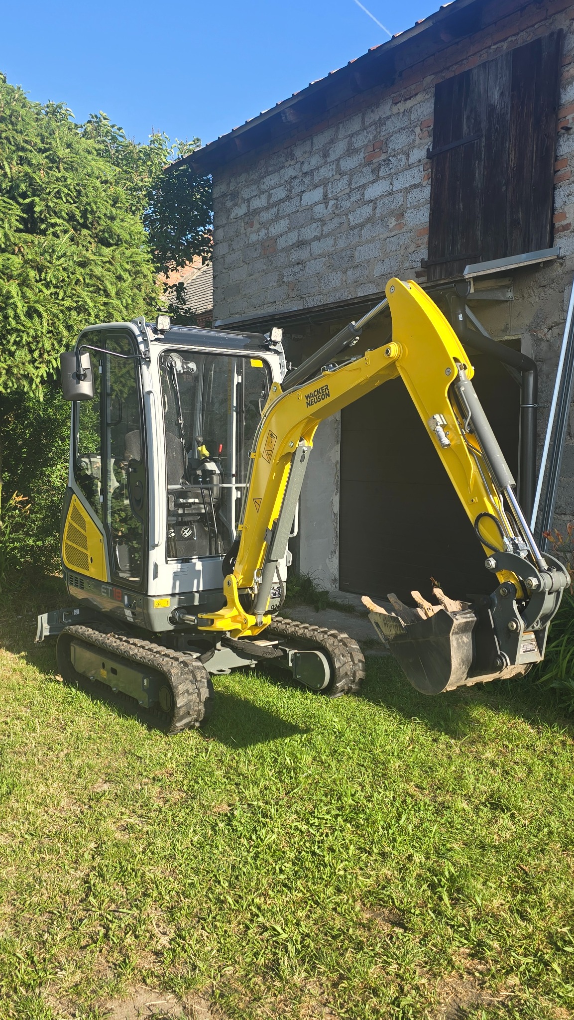 Żółto-szara mini koparka Wacker Neuson na trawie, z budynkiem w tle. Widoczne detale kabiny i łyżki. Sprzęt gotowy do pracy.