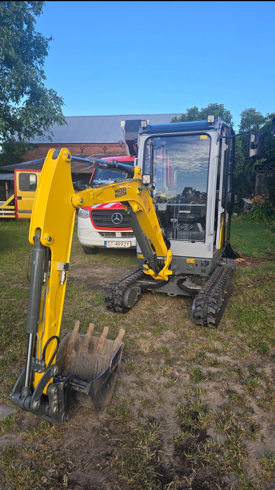 Żółto-szara mini koparka Wacker Neuson na trawiastym terenie, z widocznym łyżką i gąsienicami. W tle budynek z czerwonej cegły i biały bus z czerwonym pasem.