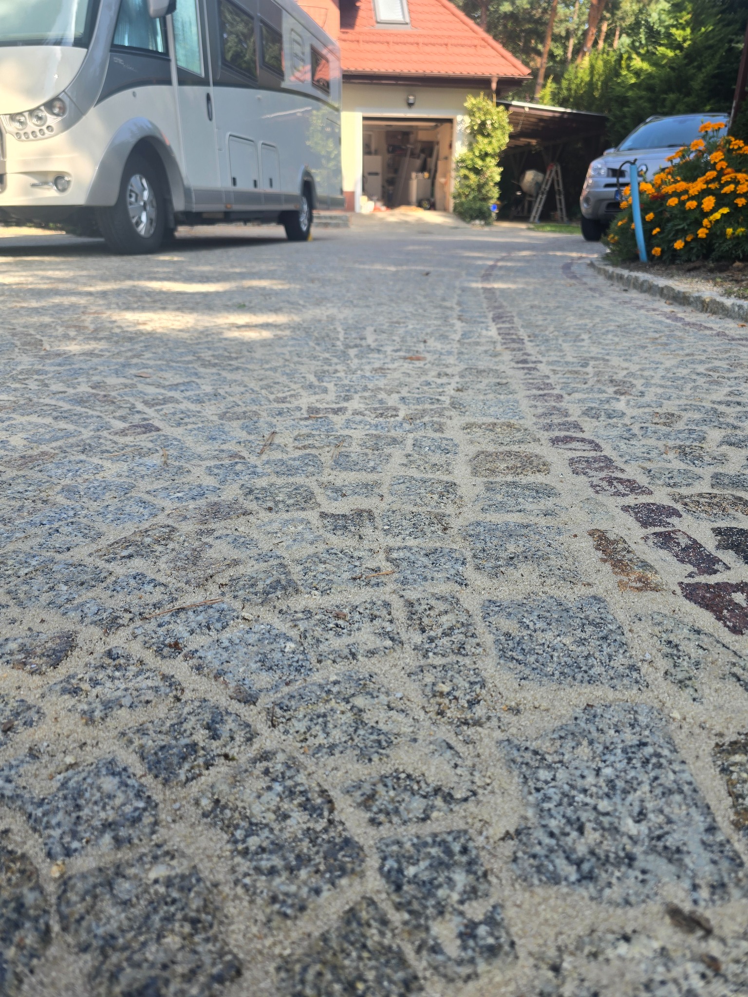 Szara kostka brukowa na podjeździe z widocznymi śladami opon, z zaparkowanym kamperem i samochodem osobowym w tle, otoczona zielenią i kwiatami.