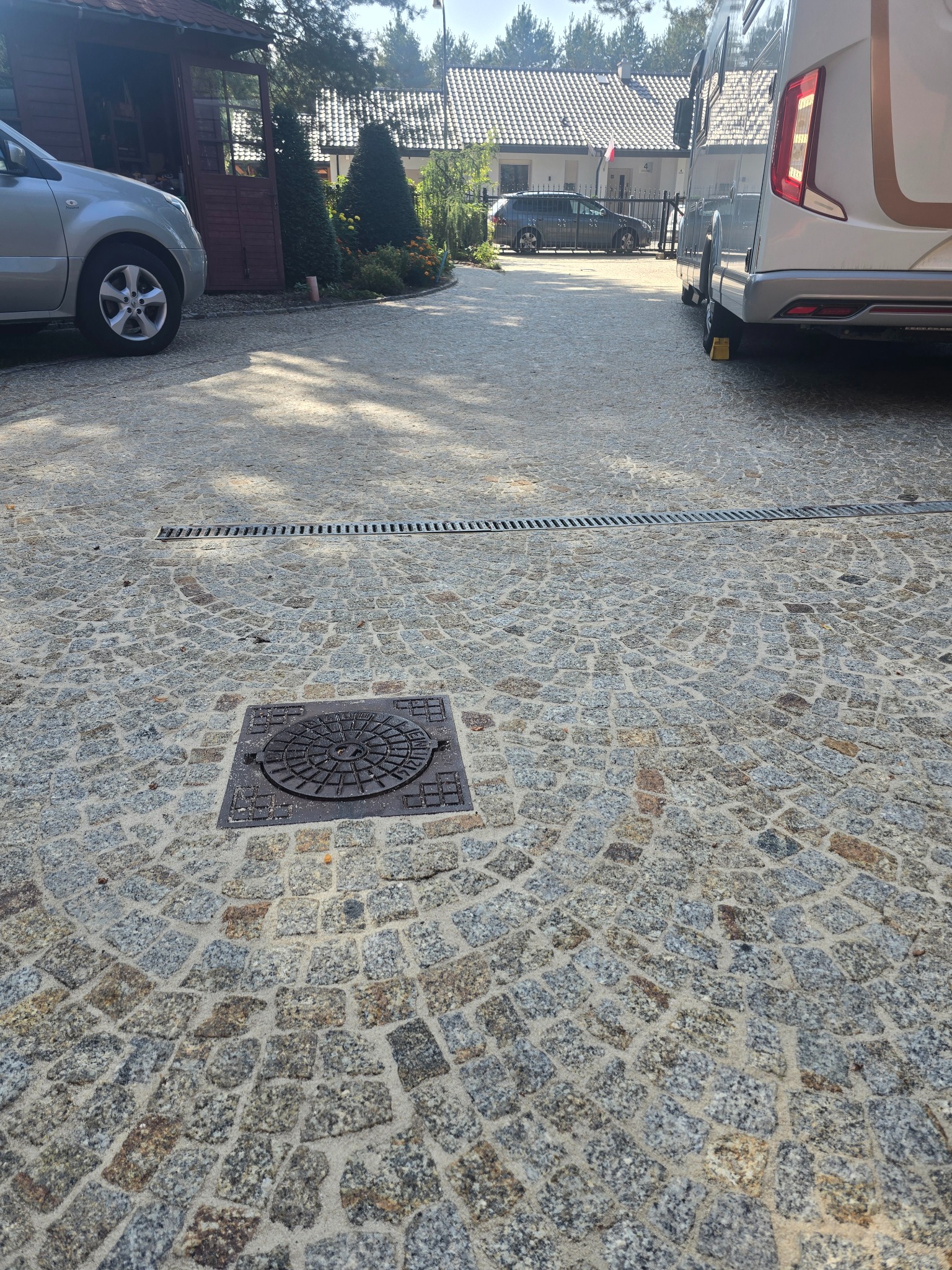 Ułożona kostka brukowa w odcieniach szarości i brązu z widoczną studzienką kanalizacyjną i liniowym odpływem. W tle dom, samochody i przyczepa kempingowa.