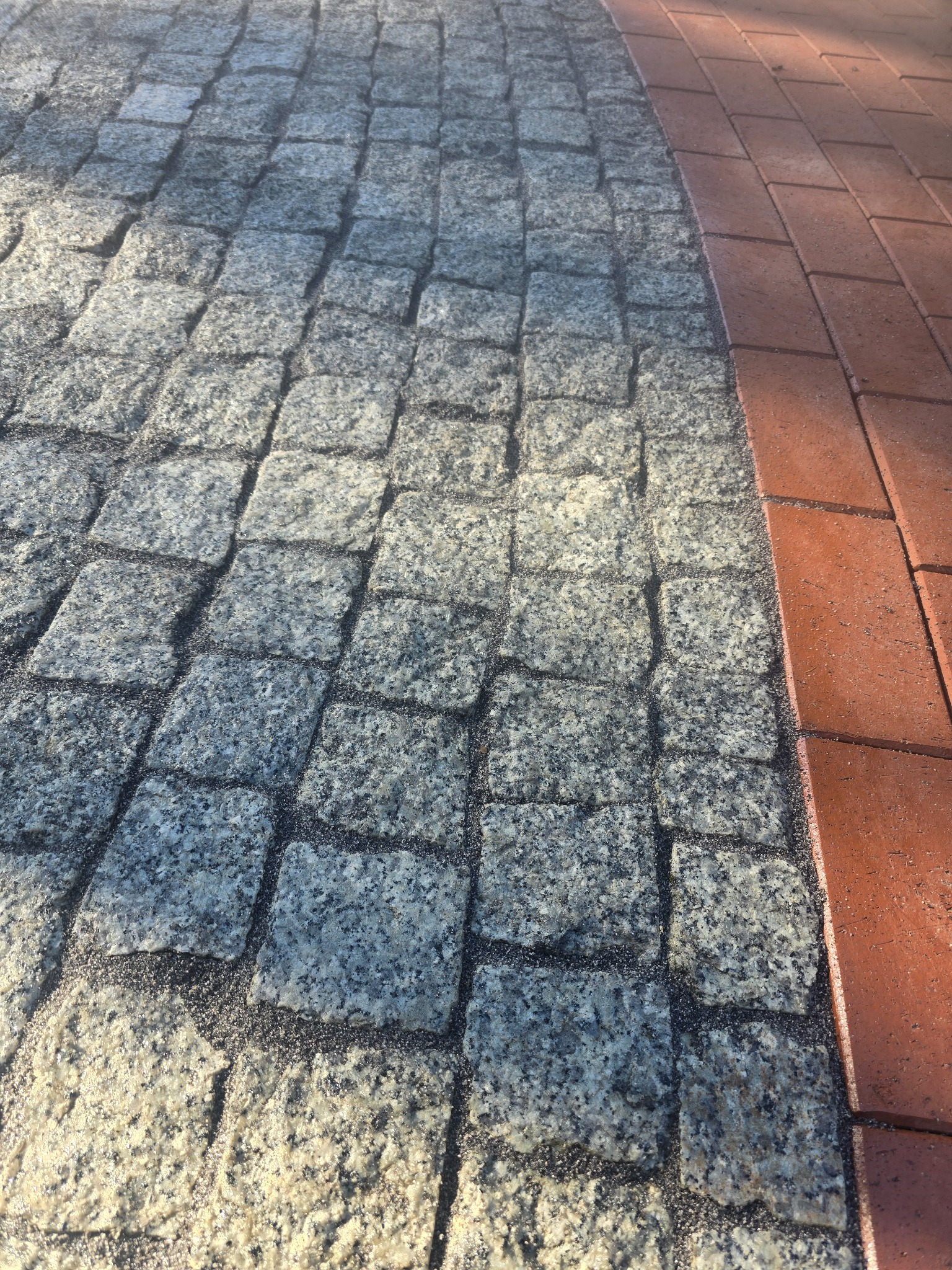 Szara kostka granitowa ułożona obok czerwonej kostki klinkierowej, tworząca kontrastową i estetyczną nawierzchnię. Widoczne drobne ziarna piasku w fugach.