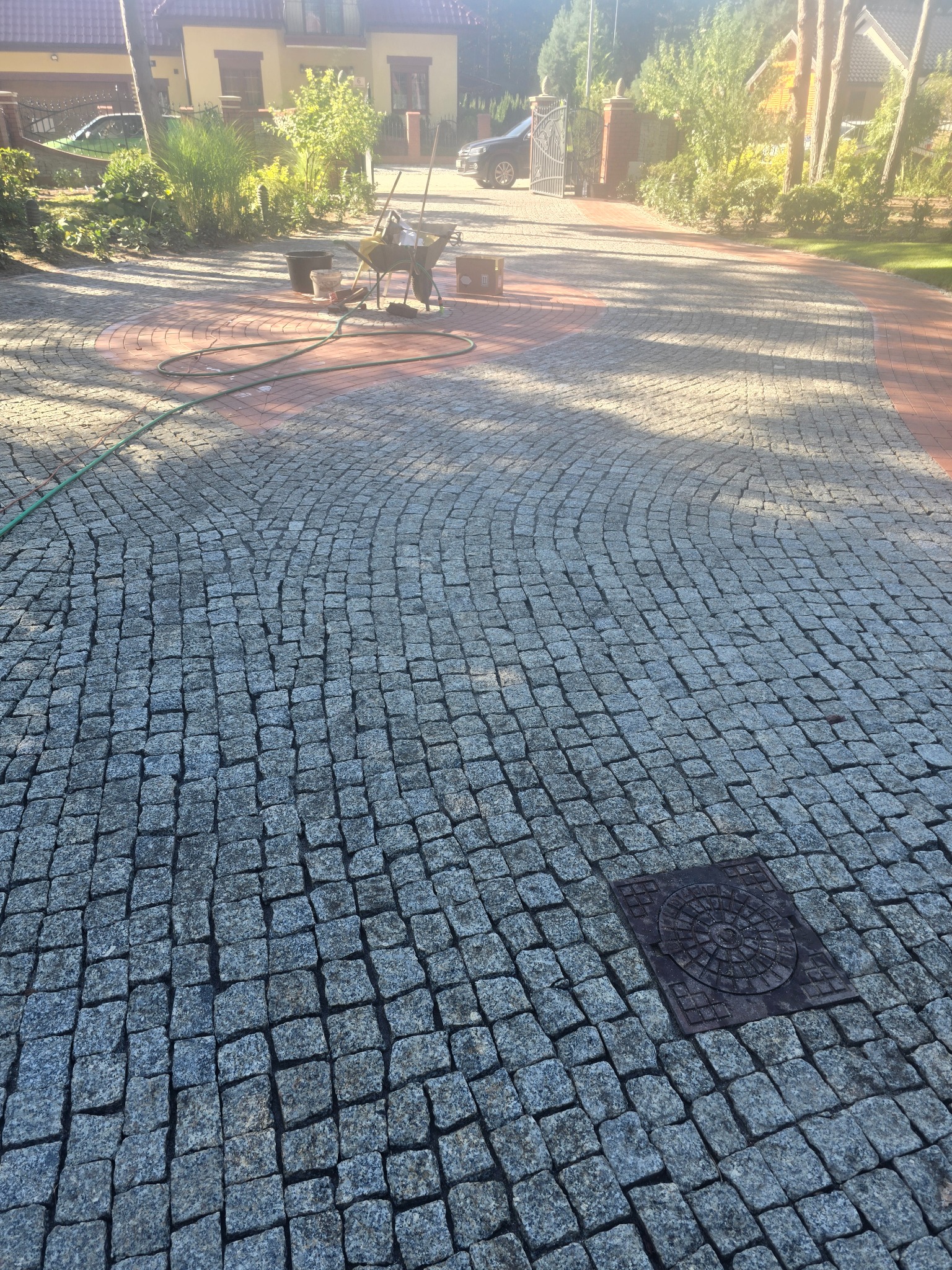 Ułożona kostka brukowa w szaro-czerwonym odcieniu, z widocznymi narzędziami pracy i wózkiem na tle domów i zieleni. Widok na posesję z bramą w tle.