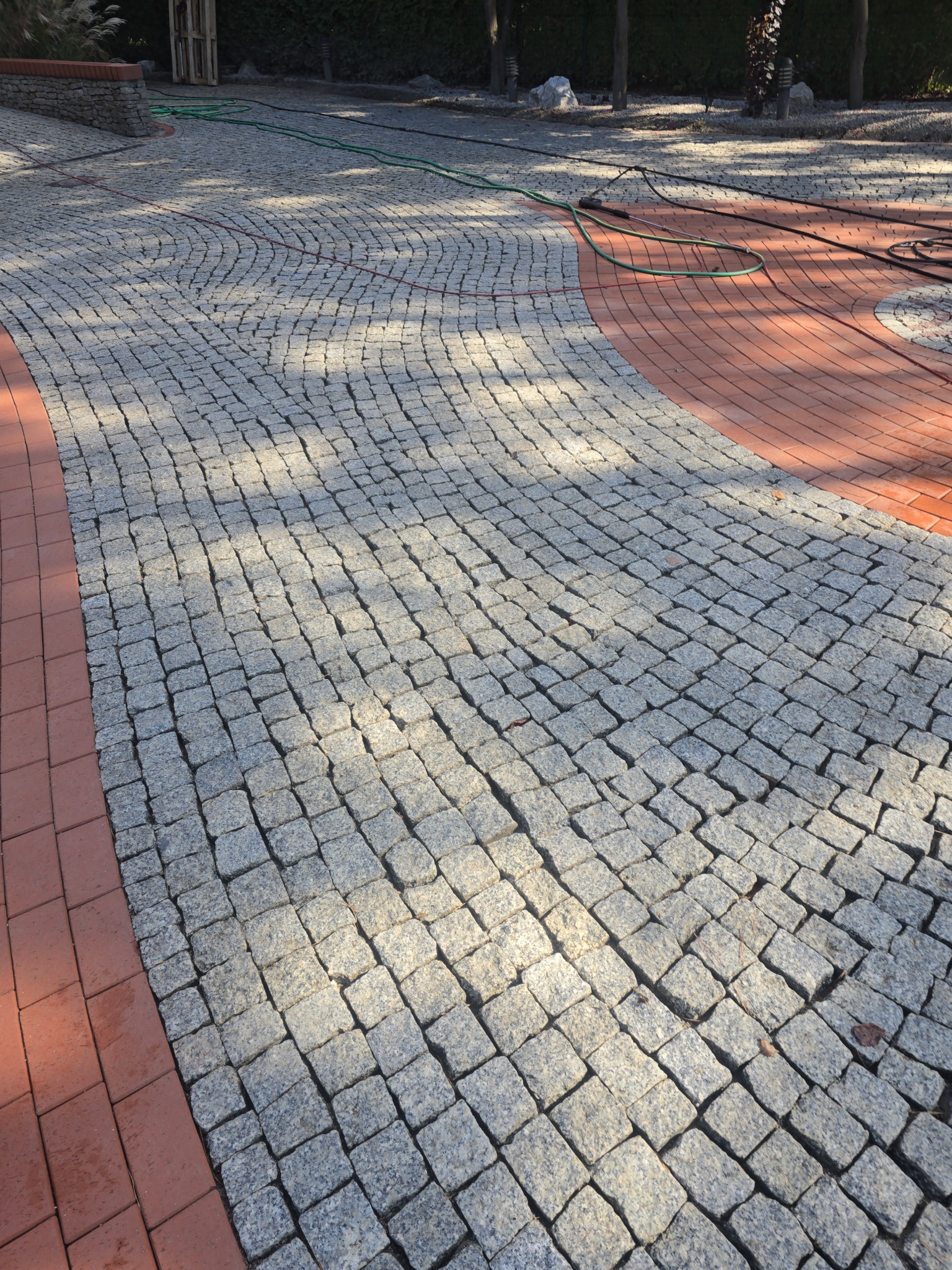 Ułożona kostka brukowa w Trzebiechowie, tworząca wzór łuku z obramowaniem z czerwonej cegły. Widoczne cienie drzew i węże ogrodowe na powierzchni.