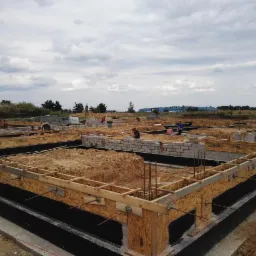 Budowa fundamentów pod dom w Świebodzicach: widok na przygotowany szalunek z deskami i zbrojenie, robotnicy w tle, pochmurne niebo.