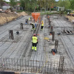 Zbrojenie fundamentów na budowie, widoczne stalowe pręty tworzące siatkę, pionowe elementy zbrojeniowe, pracownicy w odblaskowych kamizelkach oraz pomarańczowy lej do betonu zawieszony na dźwigu.