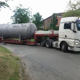 Ciężarówka MAN transportuje na niskopodwoziowej platformie duży, metalowy zbiornik ciśnieniowy w otoczeniu budynków przemysłowych i drzew.