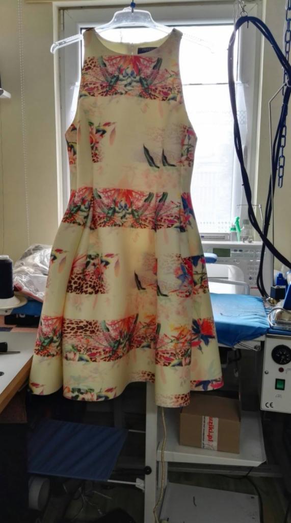 Sukienka na wieszaku z kwiatowym wzorem, w tle okno i stanowisko do prasowania z maszyną do szycia.