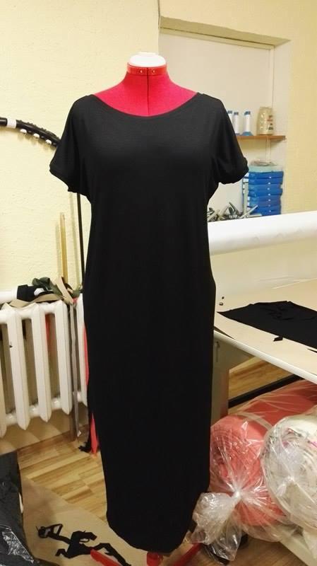 Czarna, prosta sukienka z krótkim rękawem na czerwonym manekinie krawieckim w pracowni, widoczne materiały i stół krojczy.