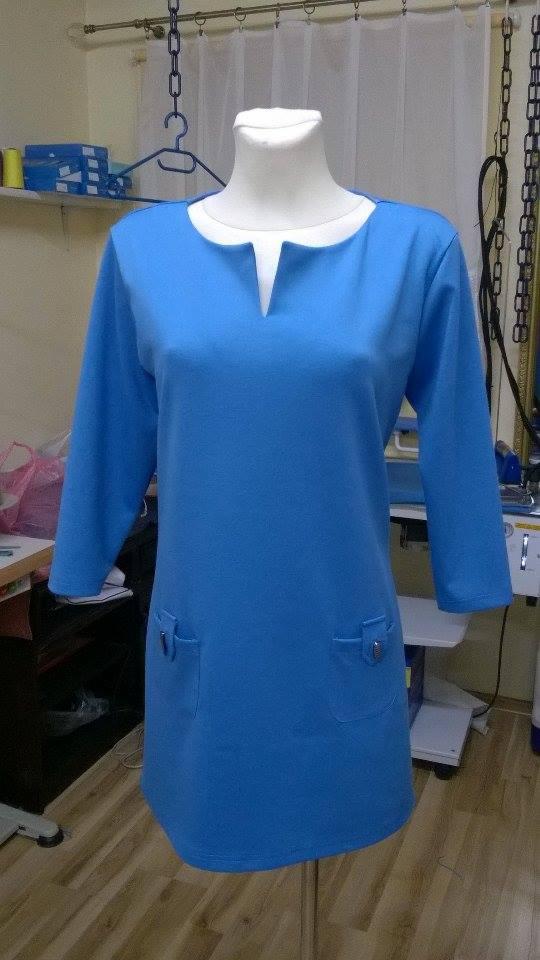 Niebieska tunika z krótkim rękawem i kieszeniami, prezentowana na manekinie w pracowni, widoczne akcesoria krawieckie w tle.