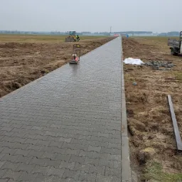 Długa ścieżka z szarej kostki brukowej ułożona na rozległym terenie, z widocznym w oddali traktorem i pracownikami, oraz ubijarką wibracyjną do zagęszczania powierzchni.