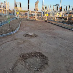 Teren przygotowany pod budowę z widocznymi wykopami, studzienką kanalizacyjną i obrzeżami chodnikowymi, na tle infrastruktury energetycznej i niebieskiego nieba.