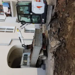 Mini koparka Bobcat E19 w Grębkowie załadowuje gruz do skrzyni samochodu ciężarowego podczas prac ziemnych w pobliżu budynku mieszkalnego.