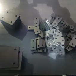 Stos kwadratowych metalowych płytek z czterema otworami obok stosu prostokątnych metalowych elementów z otworami, leżących na metalowej powierzchni.
