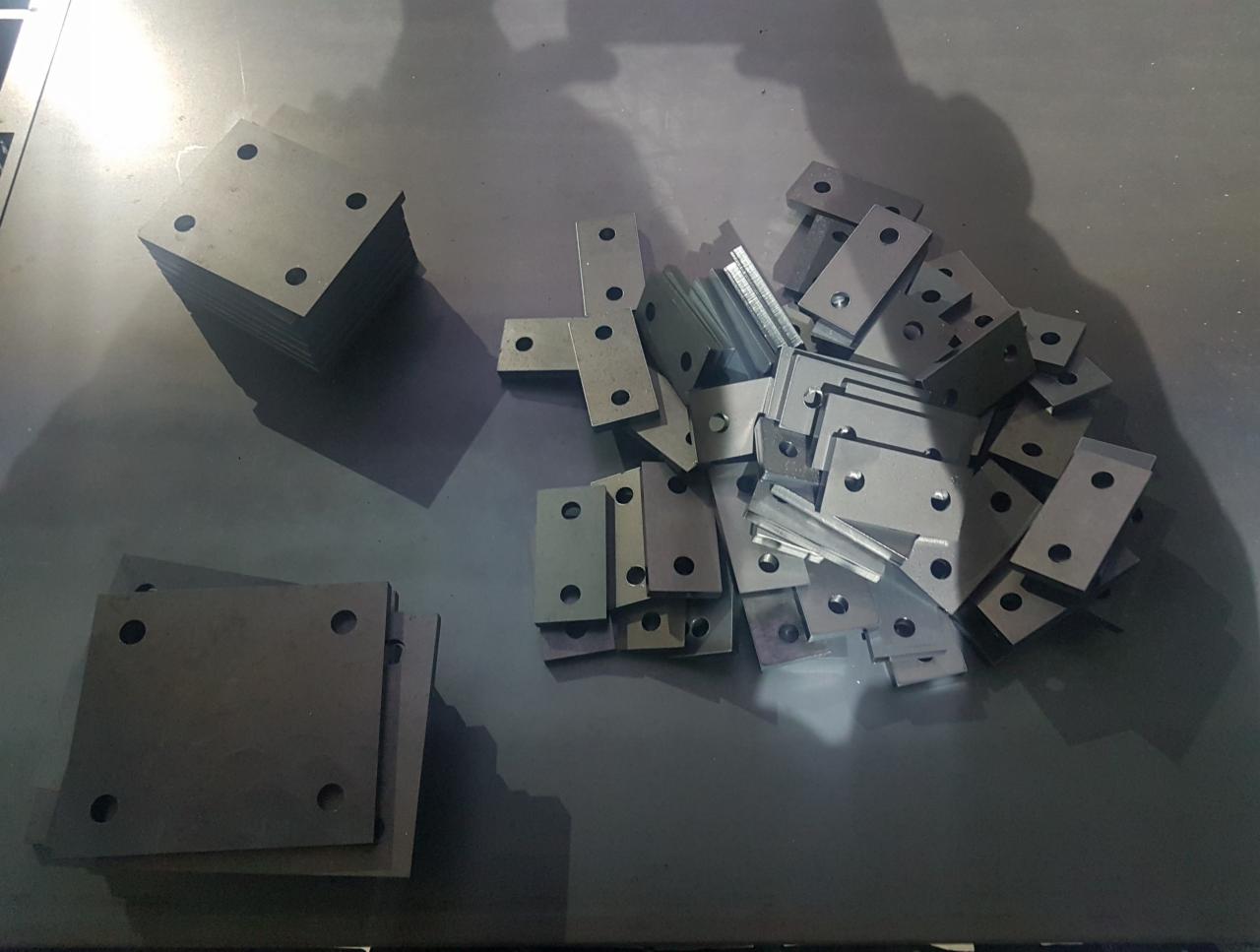 Stos kwadratowych metalowych płytek z czterema otworami obok stosu prostokątnych metalowych elementów z otworami, leżących na metalowej powierzchni.