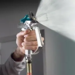 Pracownik w masce ochronnej aplikuje natryskowo gładź na ścianę za pomocą pistoletu pneumatycznego.
