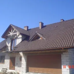 Dom w trakcie budowy z brązowym dachem z dachówki, widoczne pustaki, okna i brama garażowa, w tle drzewa i błękitne niebo.