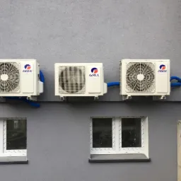 Trzy białe klimatyzatory marki Gree zamontowane na szarej ścianie budynku nad oknami z białymi ramami, z widocznymi niebieskimi rurami odprowadzającymi skropliny.