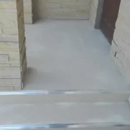 Jasnoszara, jednolita powierzchnia posadzki na schodach wejściowych z aluminiowymi listwami wykończeniowymi, kolumna z okładziną imitującą kamień na pierwszym planie.