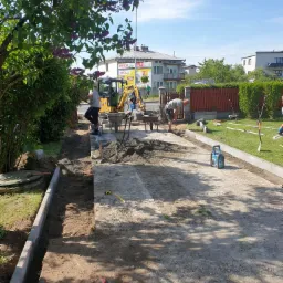 Prace przygotowawcze do ułożenia nawierzchni: wyrównywanie terenu, ustawianie obrzeży, mała koparka w tle, widoczny poziomica i inne narzędzia.