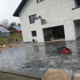 Nowo ułożona, szara kostka brukowa na podjeździe przed domem z garażem, zagęszczarka w tle, widoczne przygotowania do dalszych prac.