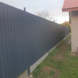 Długi, ciemnoszary, blaszany płot panelowy z betonową podmurówką, widok z boku na tle trawnika i fragmentu budynku z czerwoną dachówką.