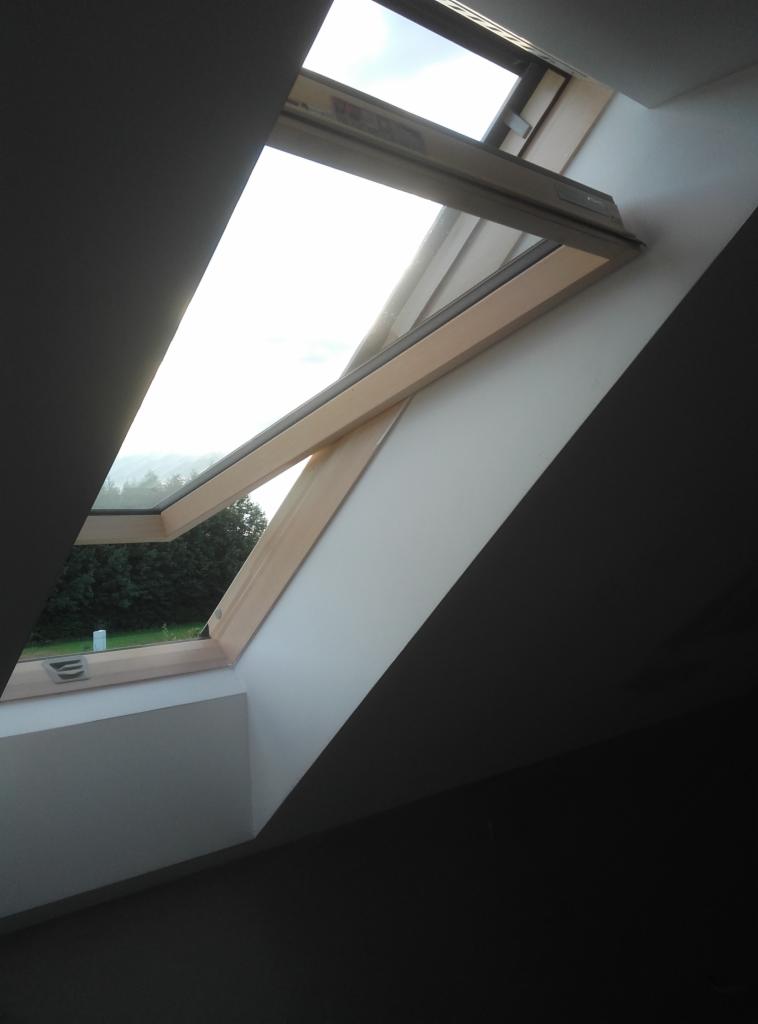 Otwarty drewniany okno dachowe w skosie poddasza, widok na zieleń na zewnątrz, świeżo pomalowane ściany w odcieniach bieli i szarości.